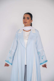 Woman wearing a baby blue abaya with tailored blazer collar and embroidered ocean-themed sleeves, paired with white scarf and heels.