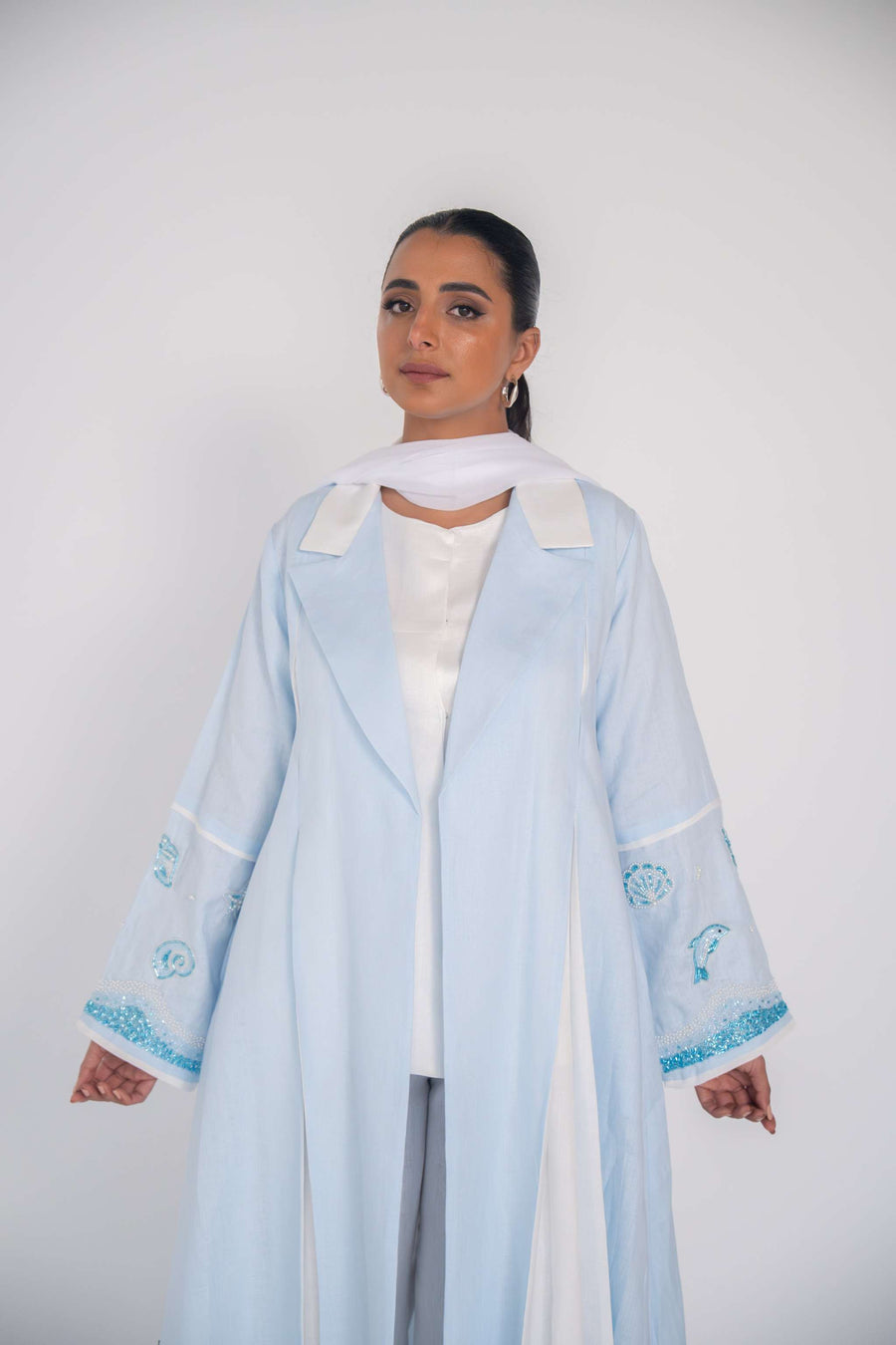 Woman wearing a baby blue abaya with tailored blazer collar and embroidered ocean-themed sleeves, paired with white scarf and heels.