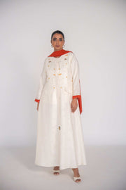 Elegant white abaya in textured jacquard fabric with hand-embroidered sunbursts and orange slice motifs, lined with vibrant orange chiffon and featuring rounded neckline and flap pockets.
