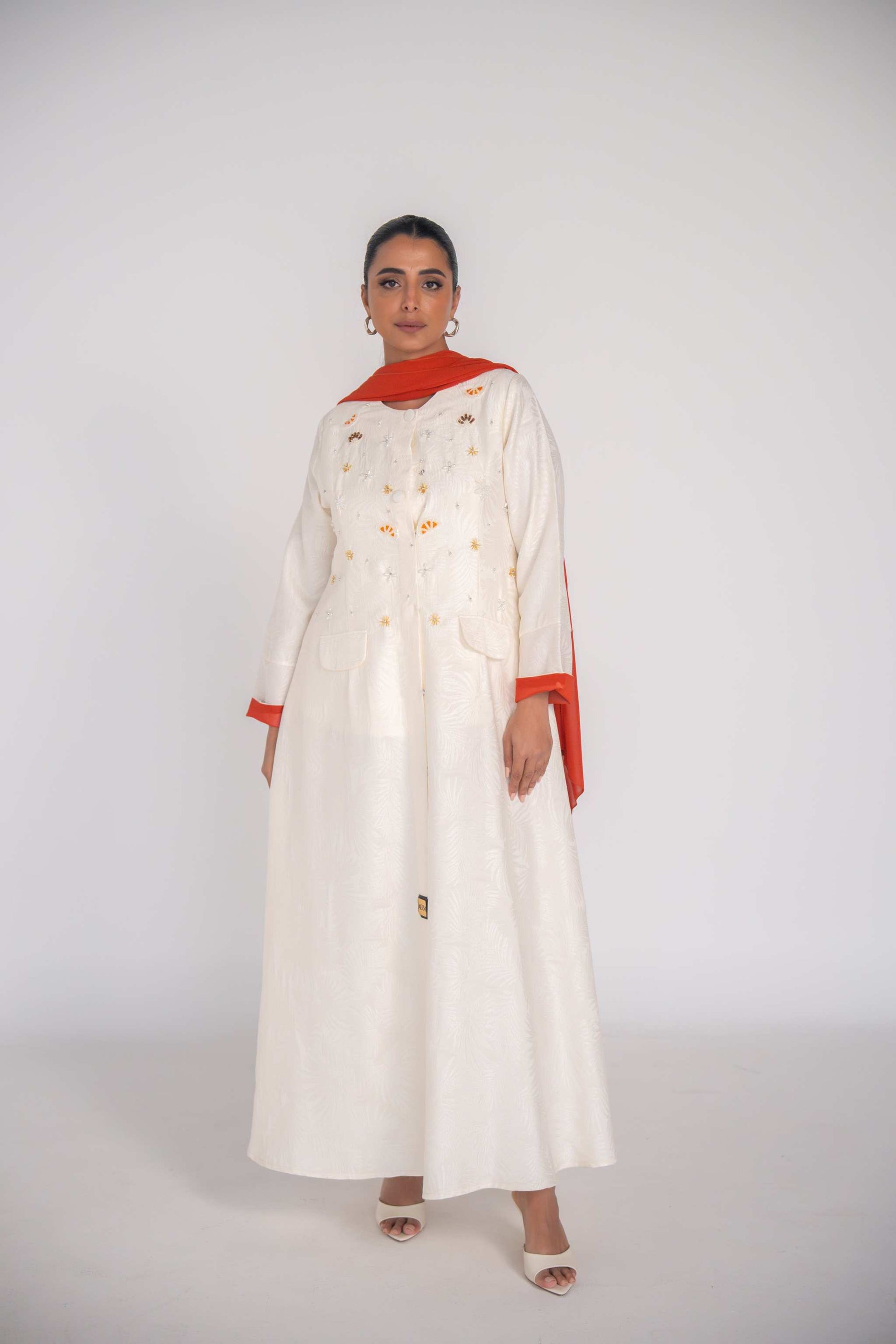 Elegant white abaya in textured jacquard fabric with hand-embroidered sunbursts and orange slice motifs, lined with vibrant orange chiffon and featuring rounded neckline and flap pockets.
