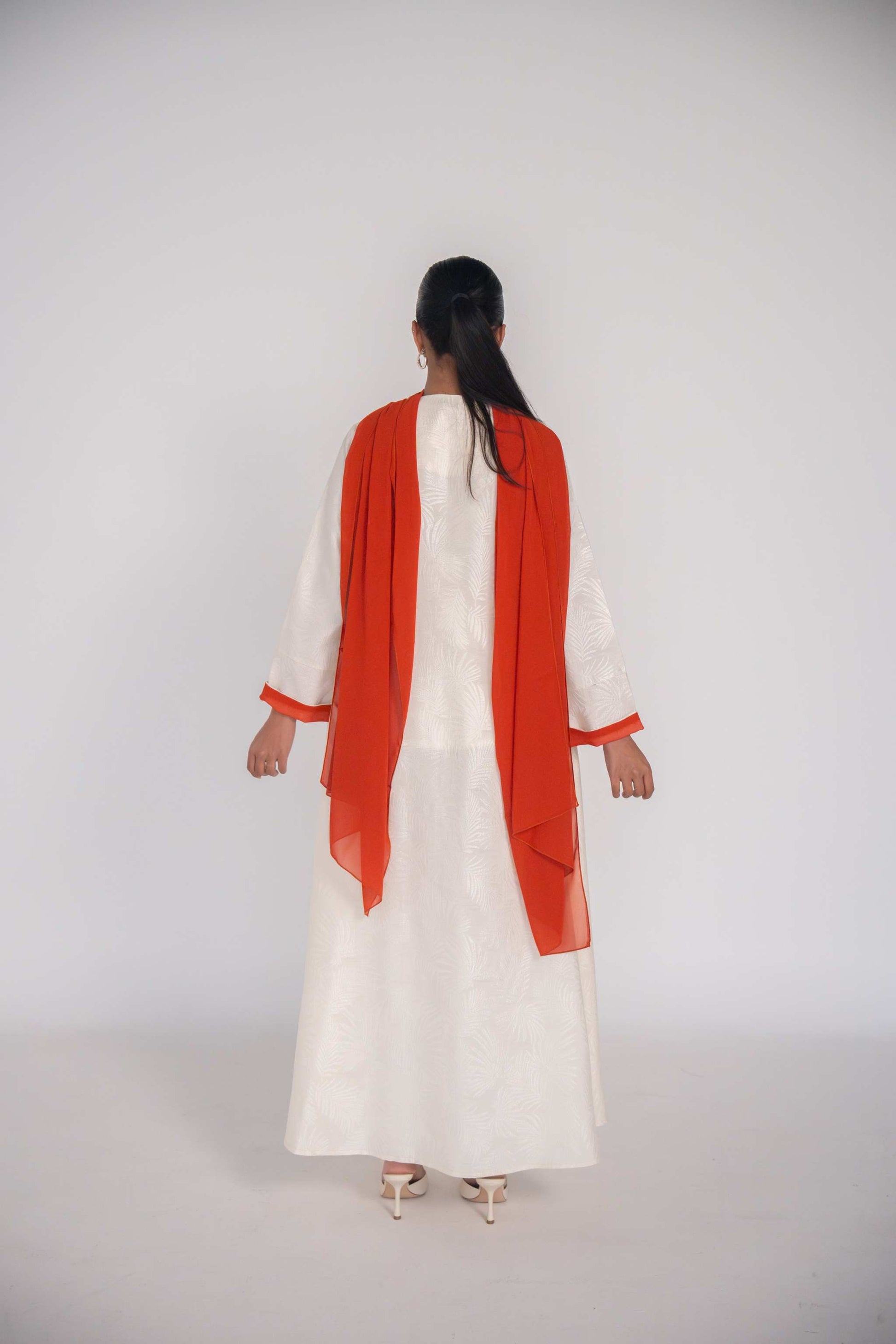 Woman wearing a white abaya made of textured jacquard fabric with an orange chiffon scarf draped over her shoulders, shown from the back.