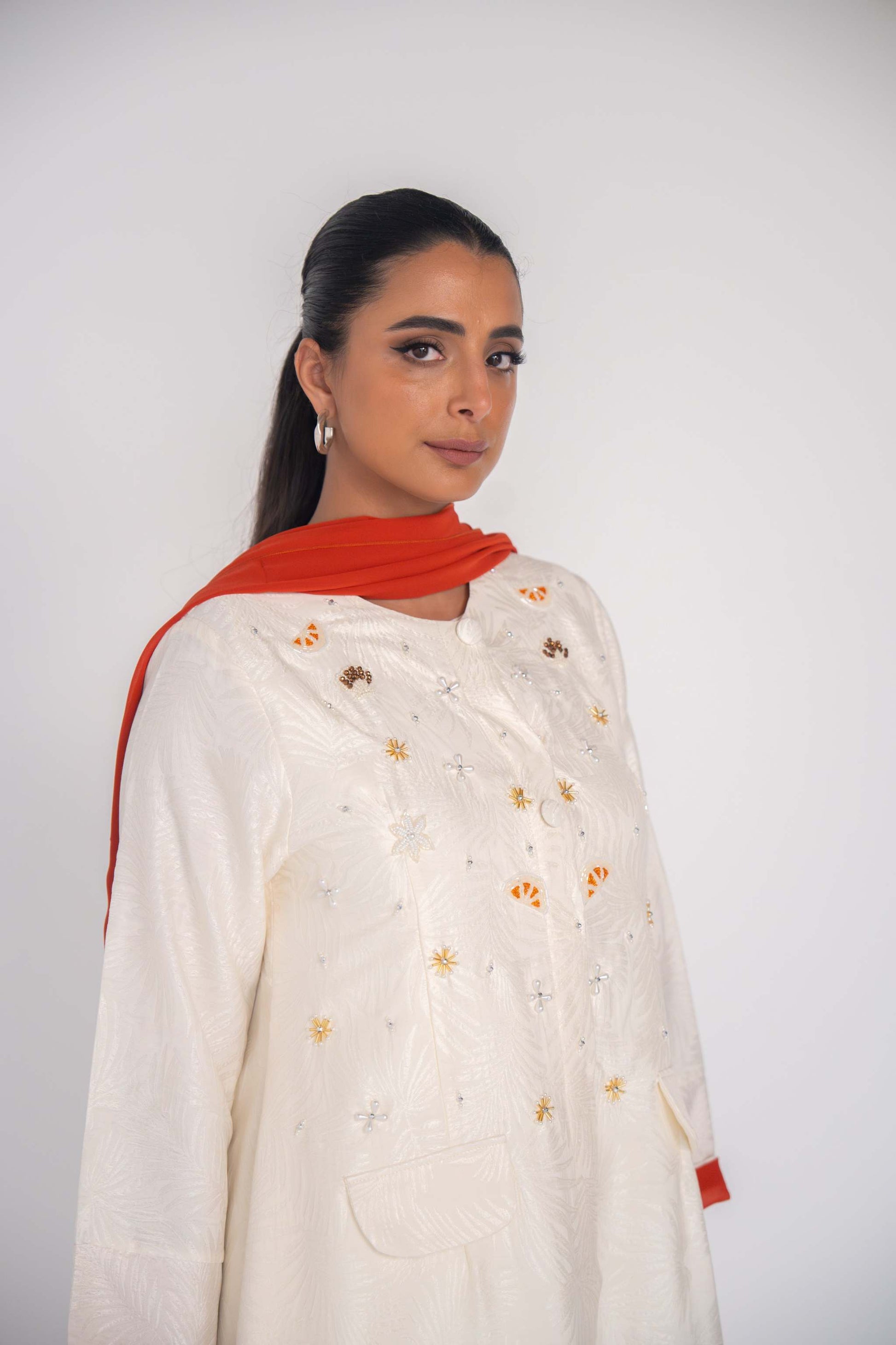 Elegant white abaya in textured jacquard fabric with hand-embroidered sunbursts and orange slice motifs, paired with a vibrant orange scarf.