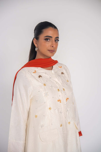 Elegant white abaya in textured jacquard fabric with hand-embroidered sunbursts and orange slice motifs, paired with a vibrant orange scarf.