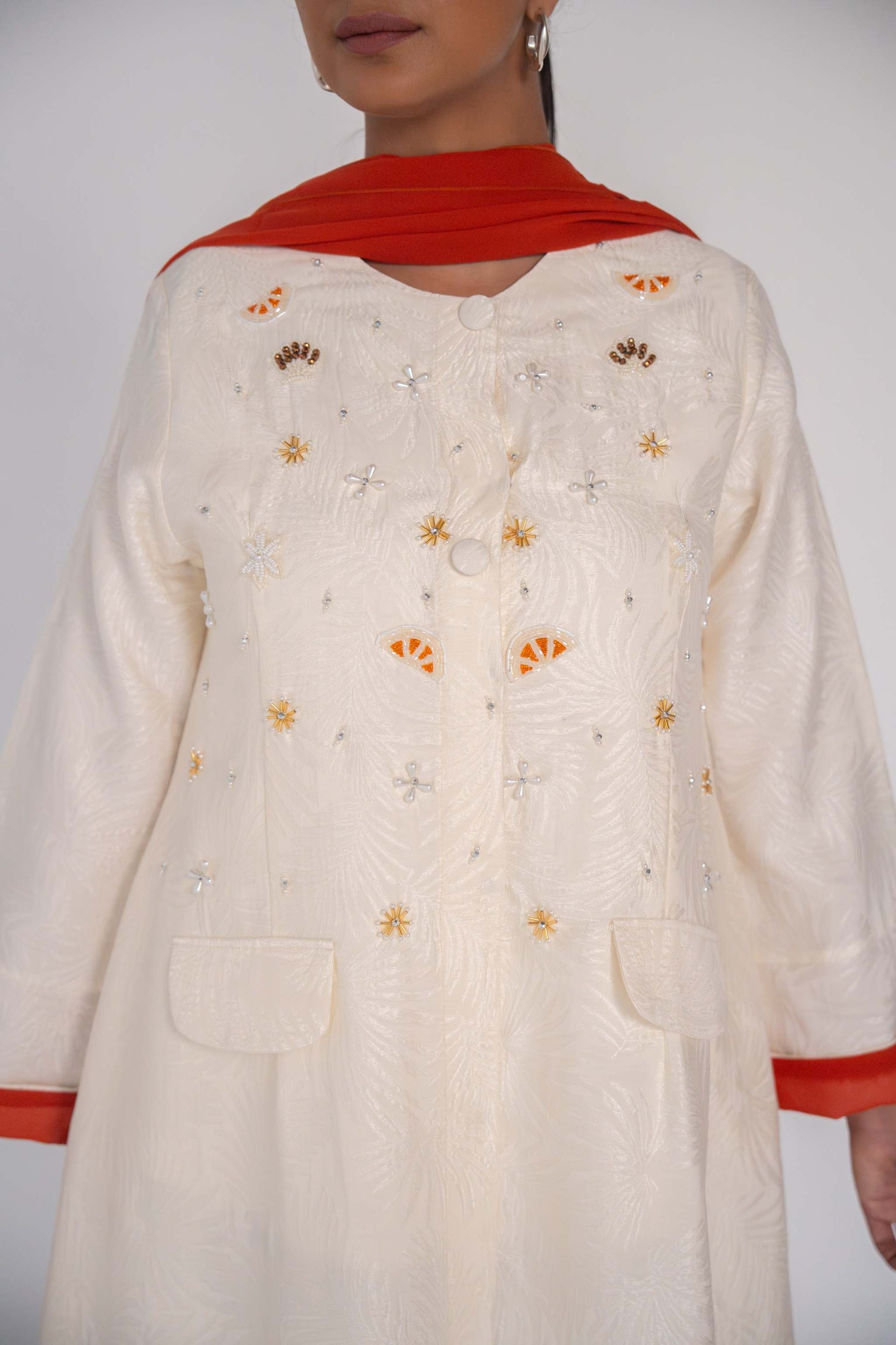 Elegant abaya in textured jacquard fabric with hand-embroidered sunbursts and orange slice motifs, accented with tonal buttons and flap pockets.