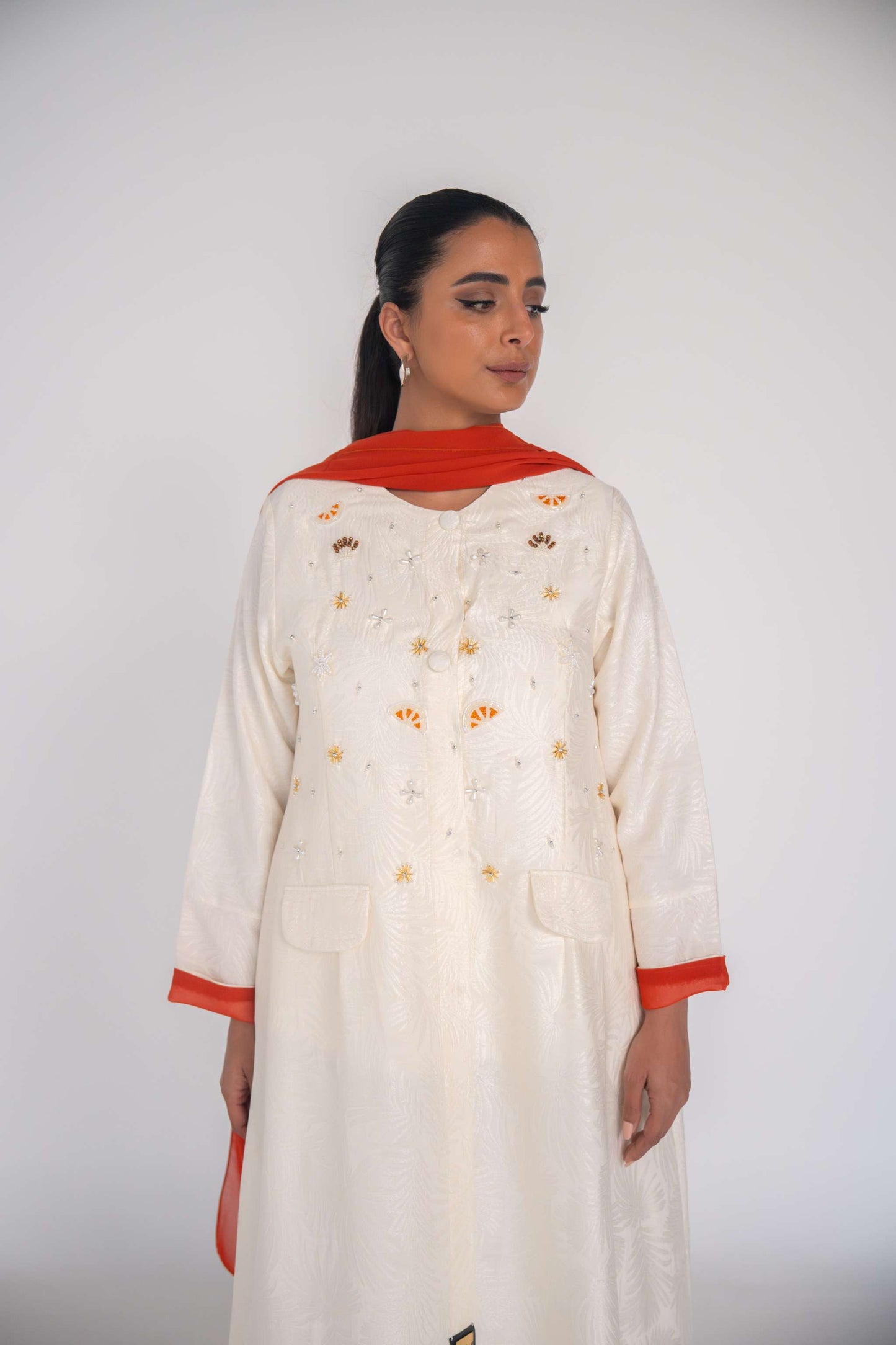 Elegant white abaya made from textured jacquard fabric with hand-embroidered sunbursts and orange slice motifs, featuring orange chiffon lining and matching scarf.