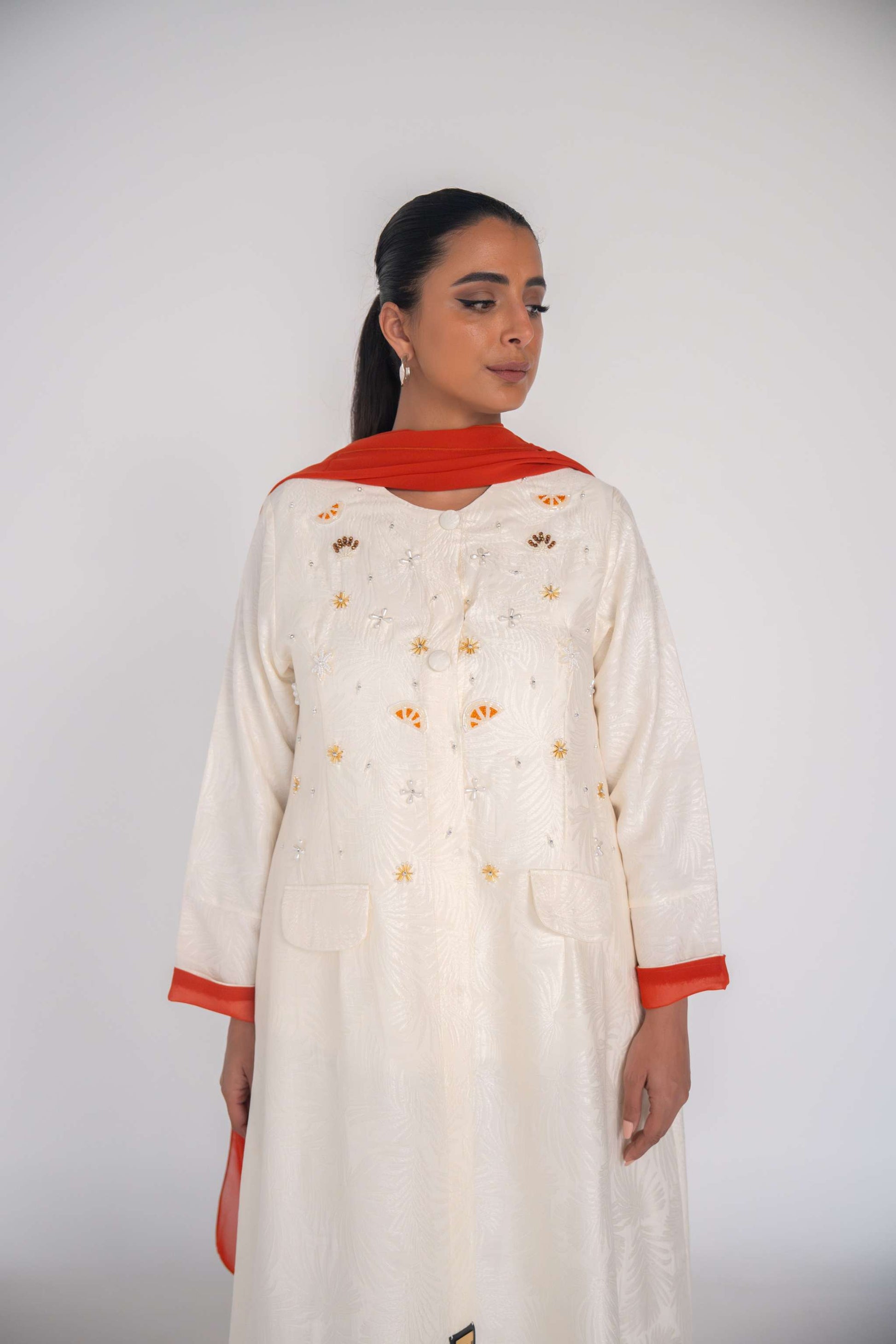 Elegant white abaya made from textured jacquard fabric with hand-embroidered sunbursts and orange slice motifs, featuring orange chiffon lining and matching scarf.