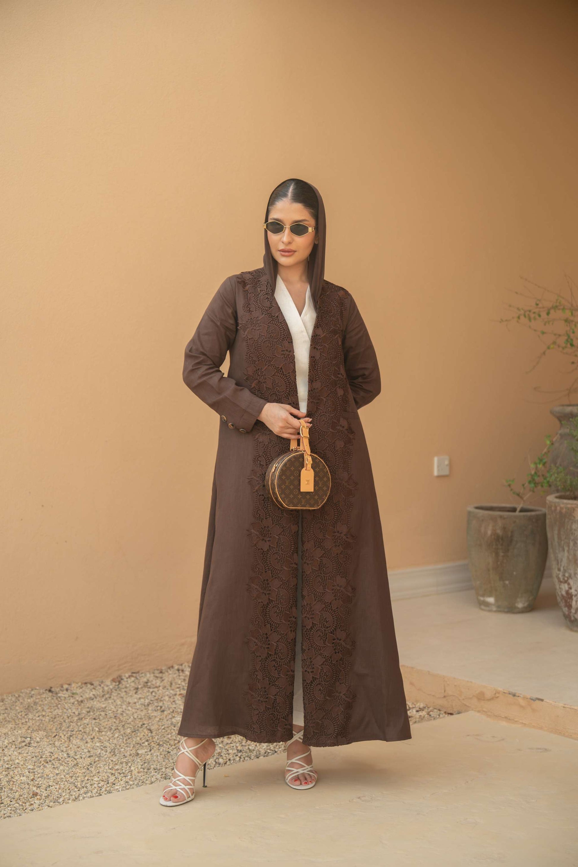 Woman wearing a deep mocha brown Mocha Bloom Abaya with floral embroidery, open front design, and sunglasses, holding a round handbag in an outdoor setting.