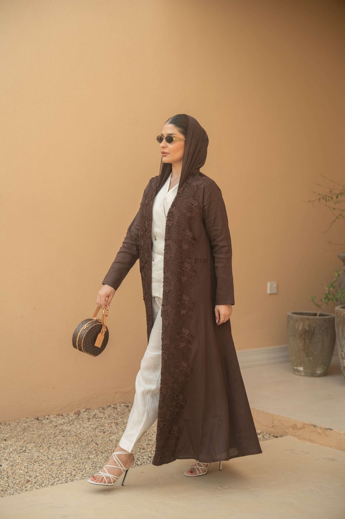 Woman wearing a deep mocha brown Mocha Bloom Abaya with floral embroidery, paired with white pants and heels, holding a round handbag.