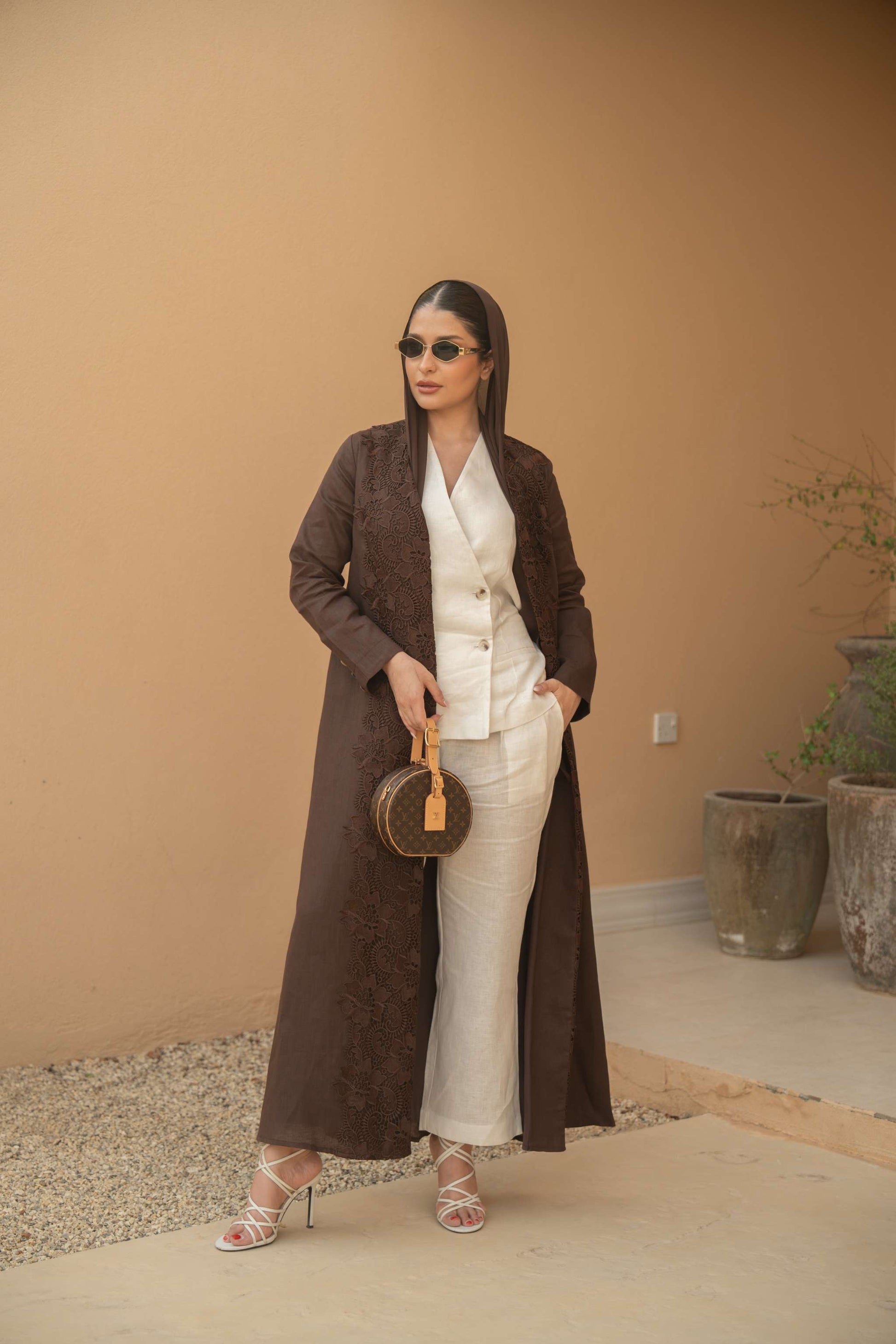 woman wearing mocha bloom abaya with floral embroidery standing against beige wall holding round handbag