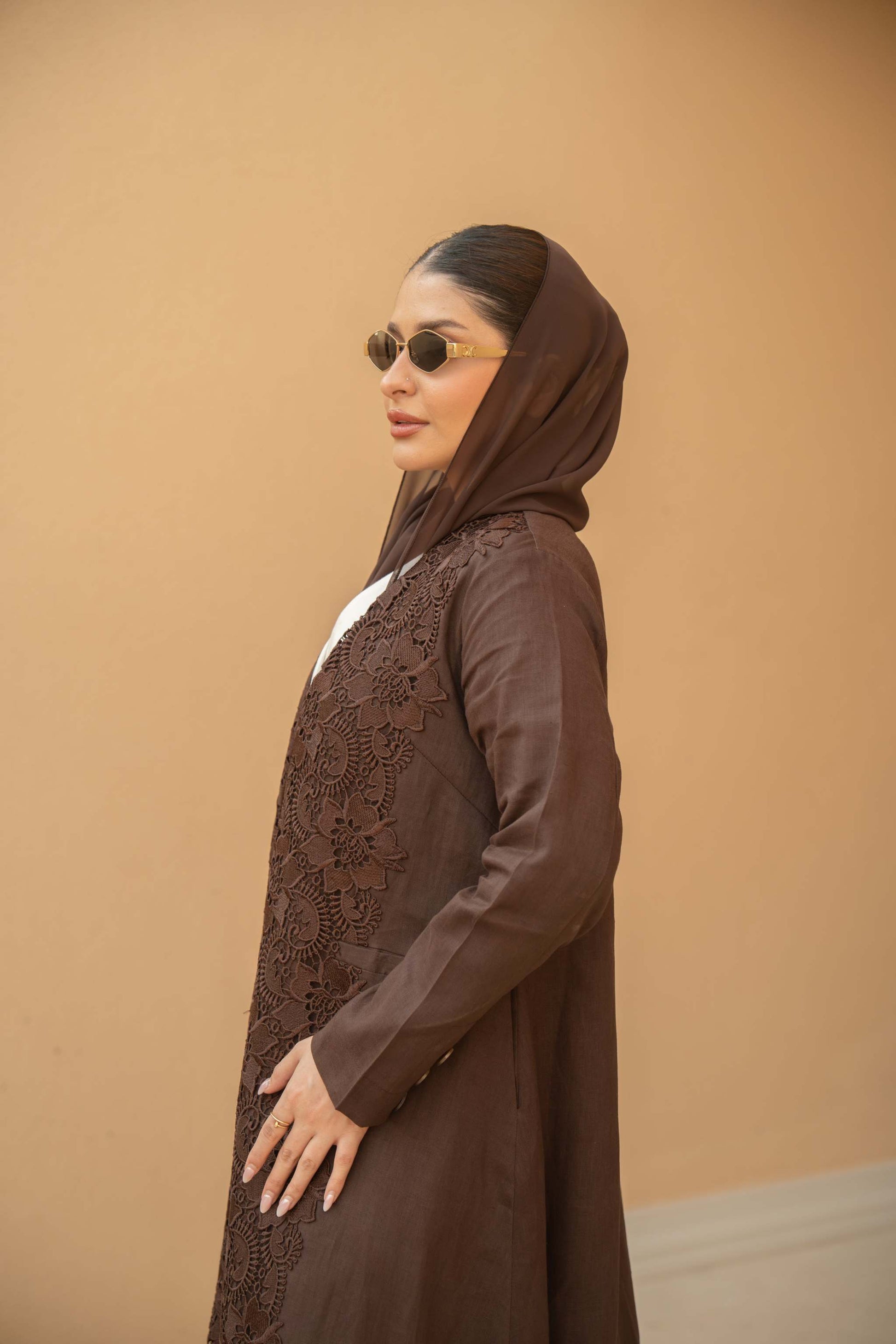 Woman wearing a deep mocha brown Mocha Bloom Abaya with floral lace embroidery and an open front design against a beige background