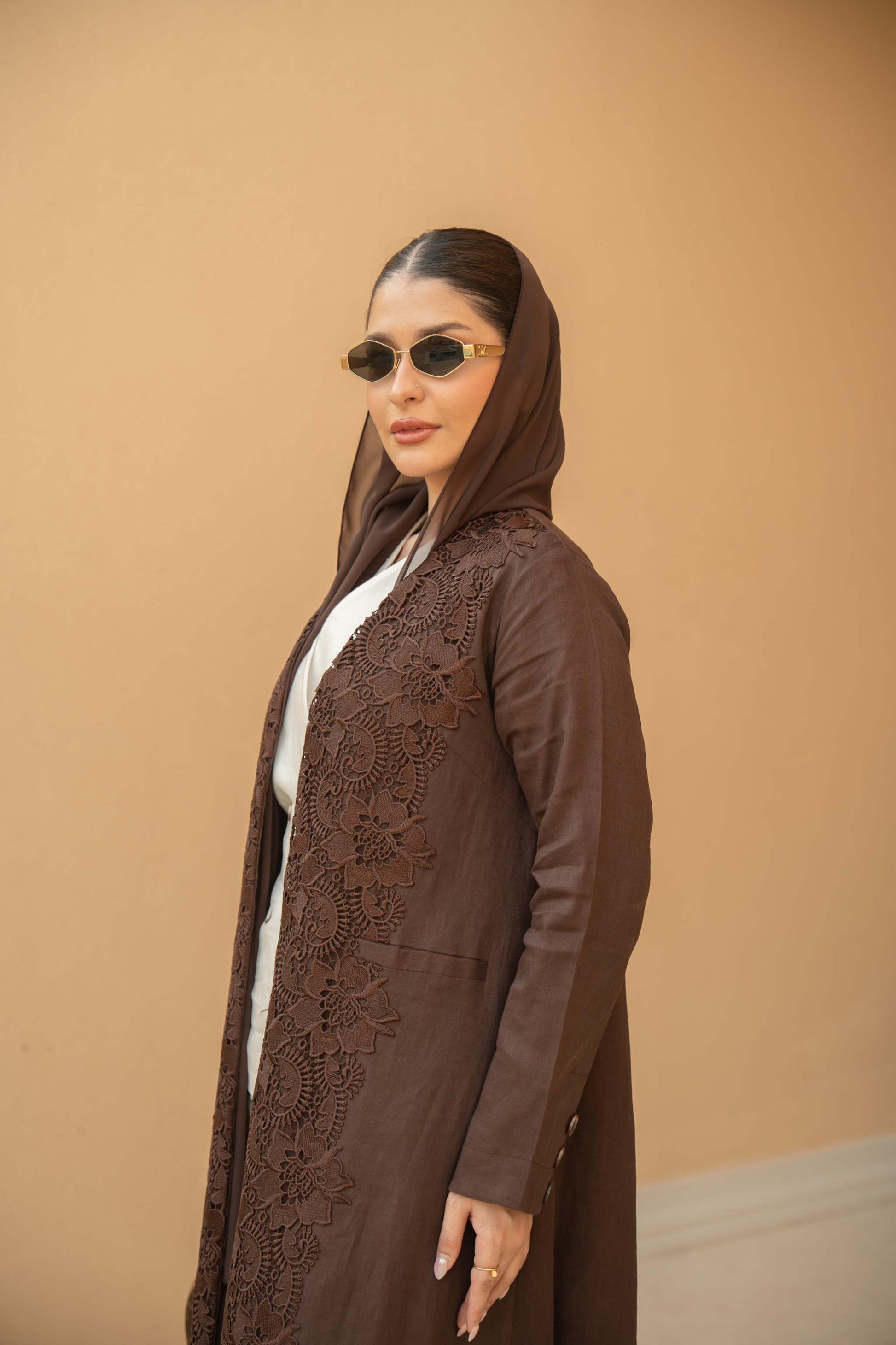 Woman wearing a deep mocha brown Mocha Bloom Abaya with floral lace embroidery and open front design against a beige background.