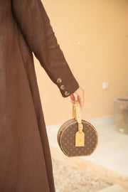 Close-up of a woman wearing a deep mocha brown Mocha Bloom Abaya holding a designer round handbag with gold accents against a neutral background