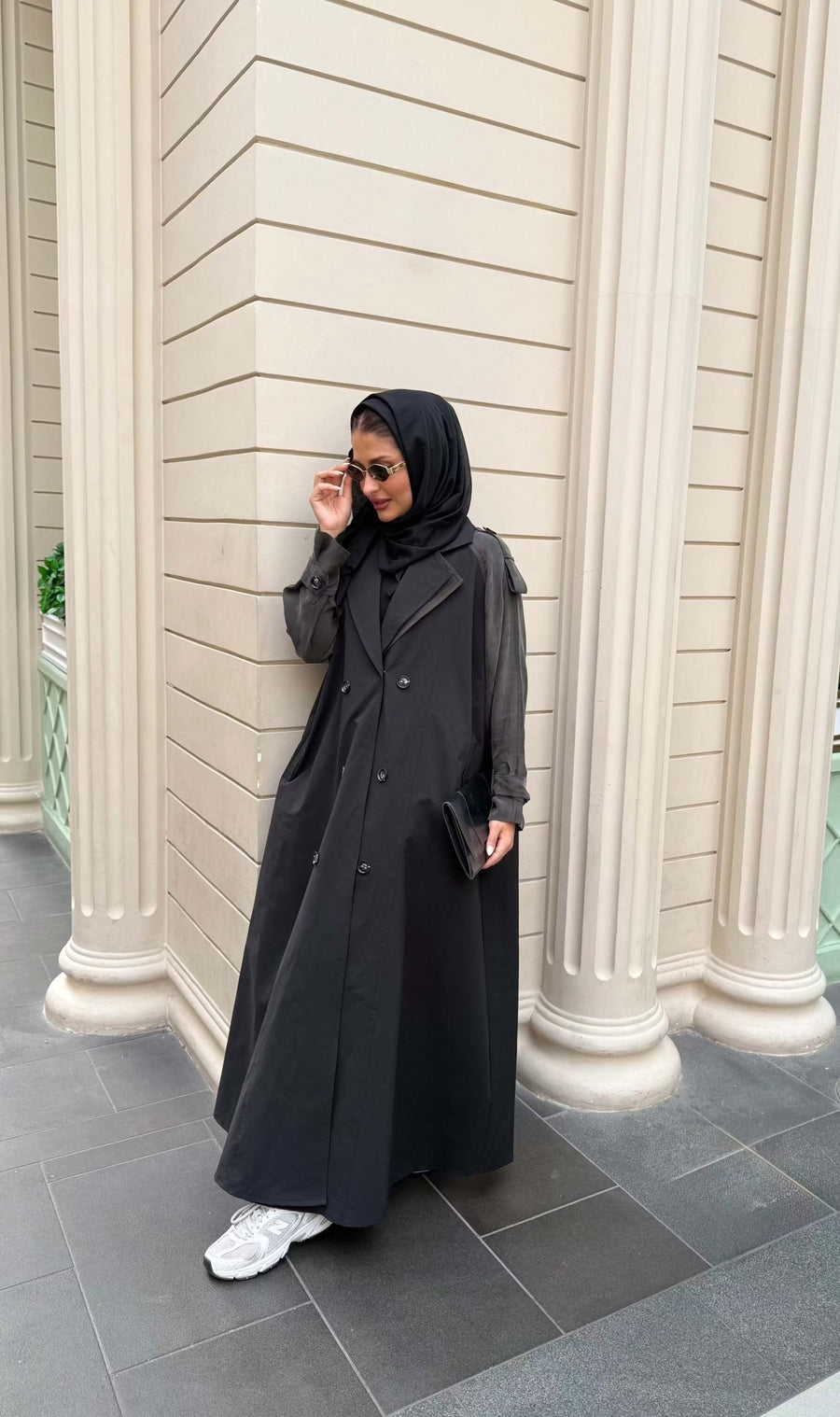 Woman wearing a black trench coat with unique sleeved design and sunglasses holding a black clutch bag - trench coat and sleeves