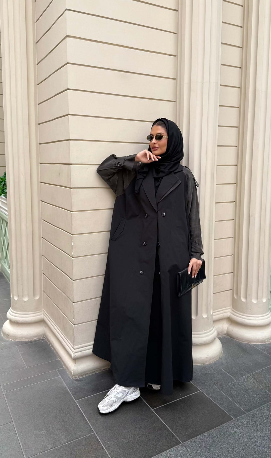 Woman wearing a black trench coat with unique sleeved design and sunglasses holding a black clutch bag - trench coat and sleeves
