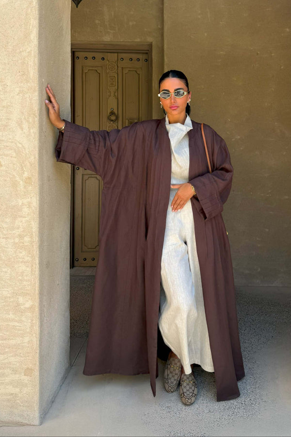 Woman wearing brown coconut linen abaya over white outfit standing in traditional beige architectural setting