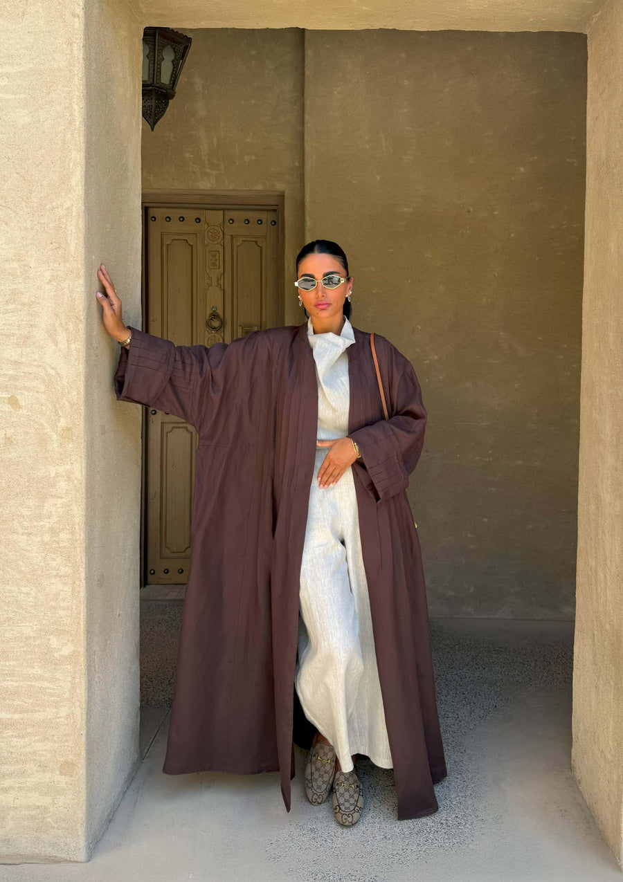 Woman wearing brown coconut linen abaya over white outfit standing in traditional beige architectural setting