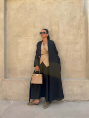 Minimalist black linen abaya with straight cut and open front worn over beige outfit with sunglasses and handbag against beige wall