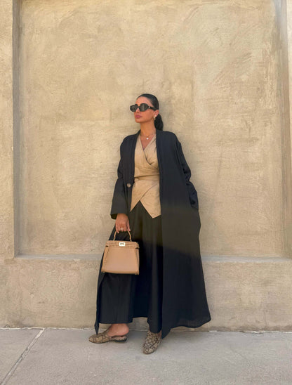Minimalist black linen abaya with straight cut and open front worn over beige outfit with sunglasses and handbag against beige wall