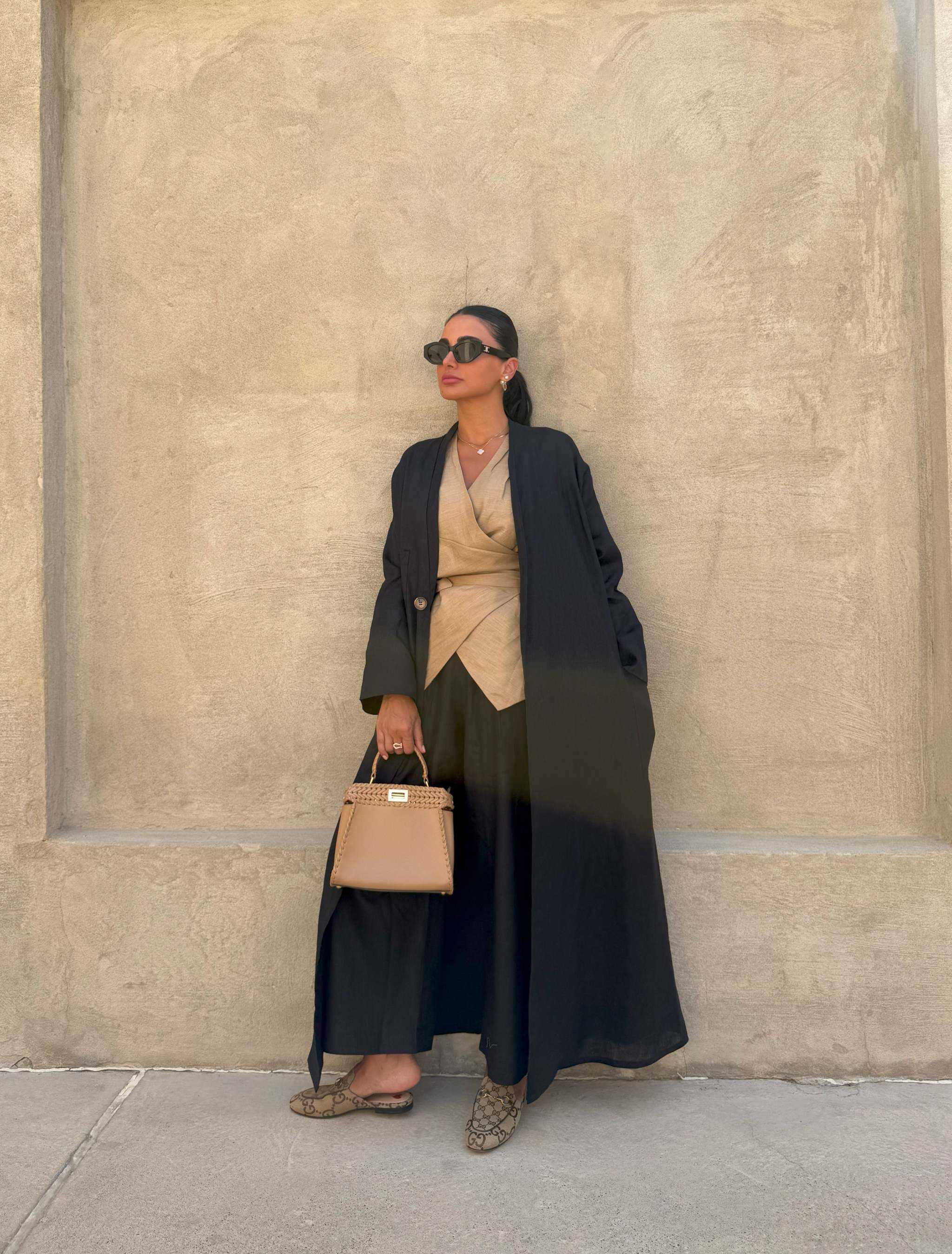 Woman wearing a lightweight black linen open-front abaya styled with beige wrap top, wide-leg pants, sunglasses, and a tan handbag against a beige wall - Midnight Linen.