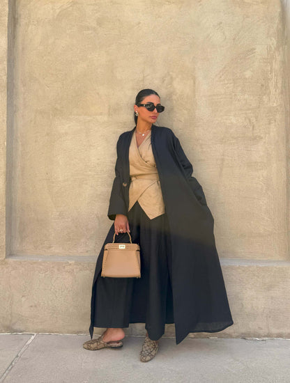 Woman wearing minimalist black linen abaya with straight cut and open front, paired with beige outfit and sunglasses against beige wall