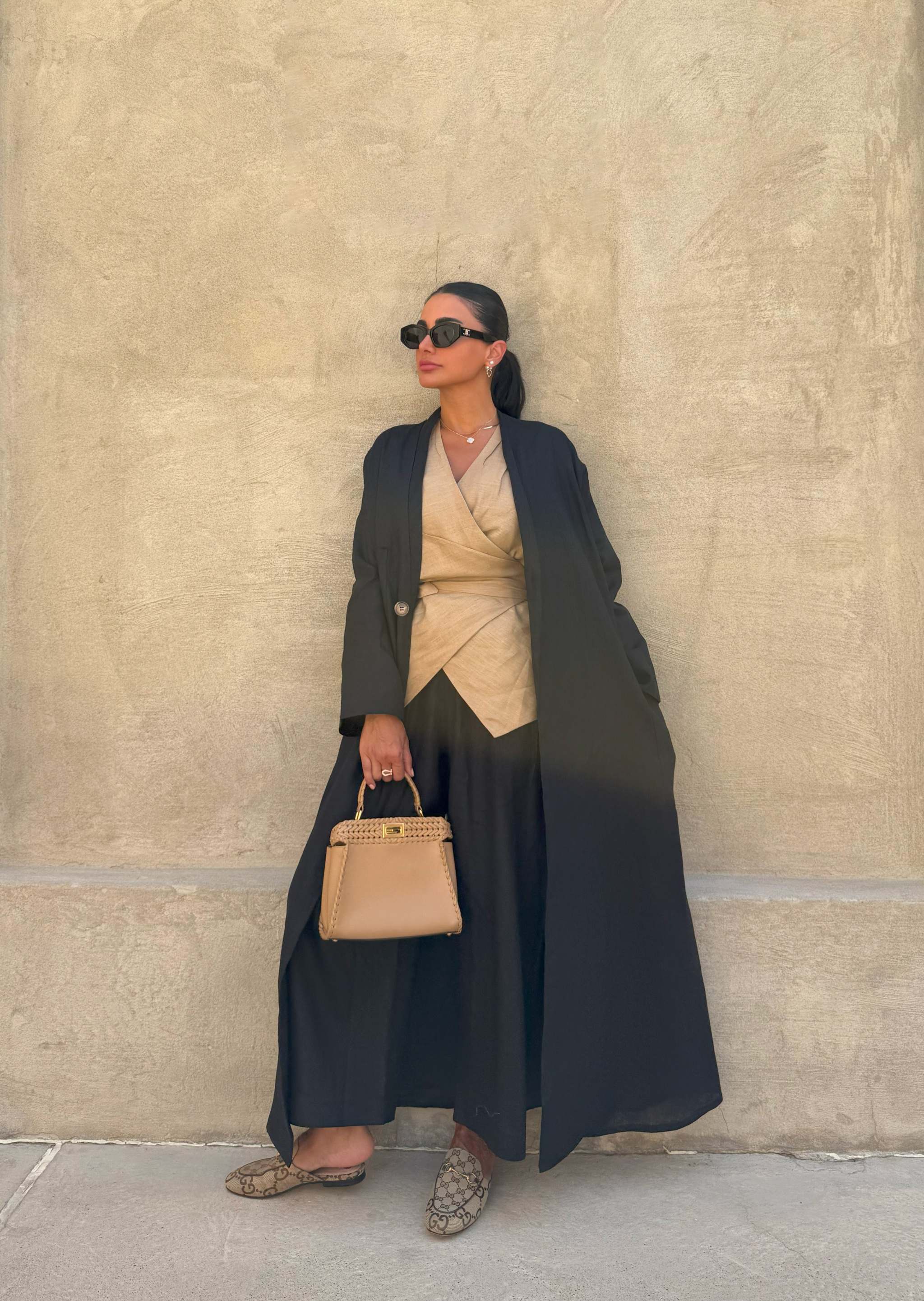 Woman wearing a lightweight black linen open-front abaya styled with beige wrap top, wide-leg pants, sunglasses, and a tan handbag against a beige wall - Midnight Linen.