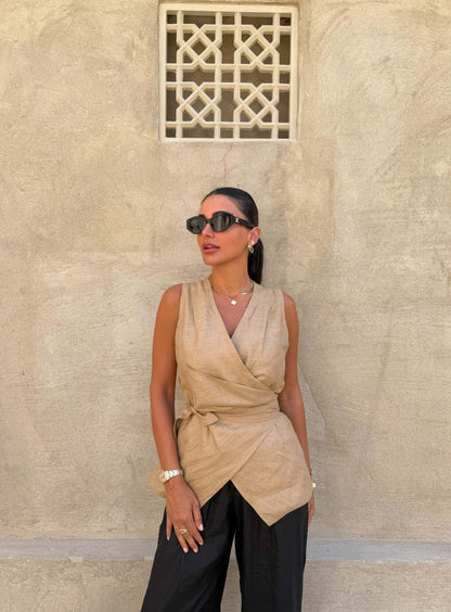 Woman wearing a sleeveless beige Clay Vest wrap top with a waist tie and black pants against a textured beige wall background