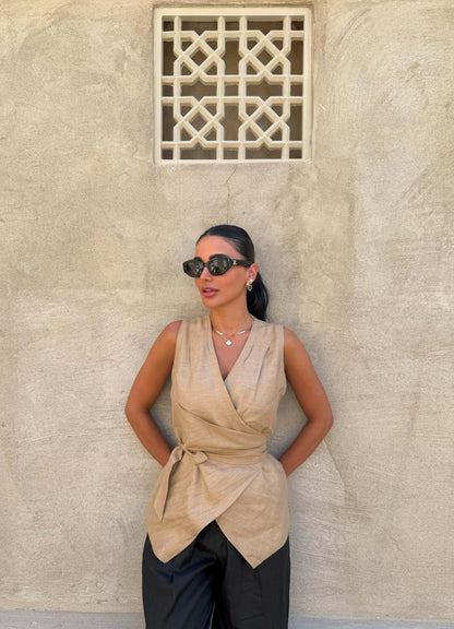 Woman wearing sleeveless beige clay vest with waist tie and black pants against textured wall background