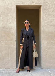 Woman wearing Latte Smoke trench-style black abaya with beige side panels and cuffs, double-breasted design and structured collar, holding beige handbag.