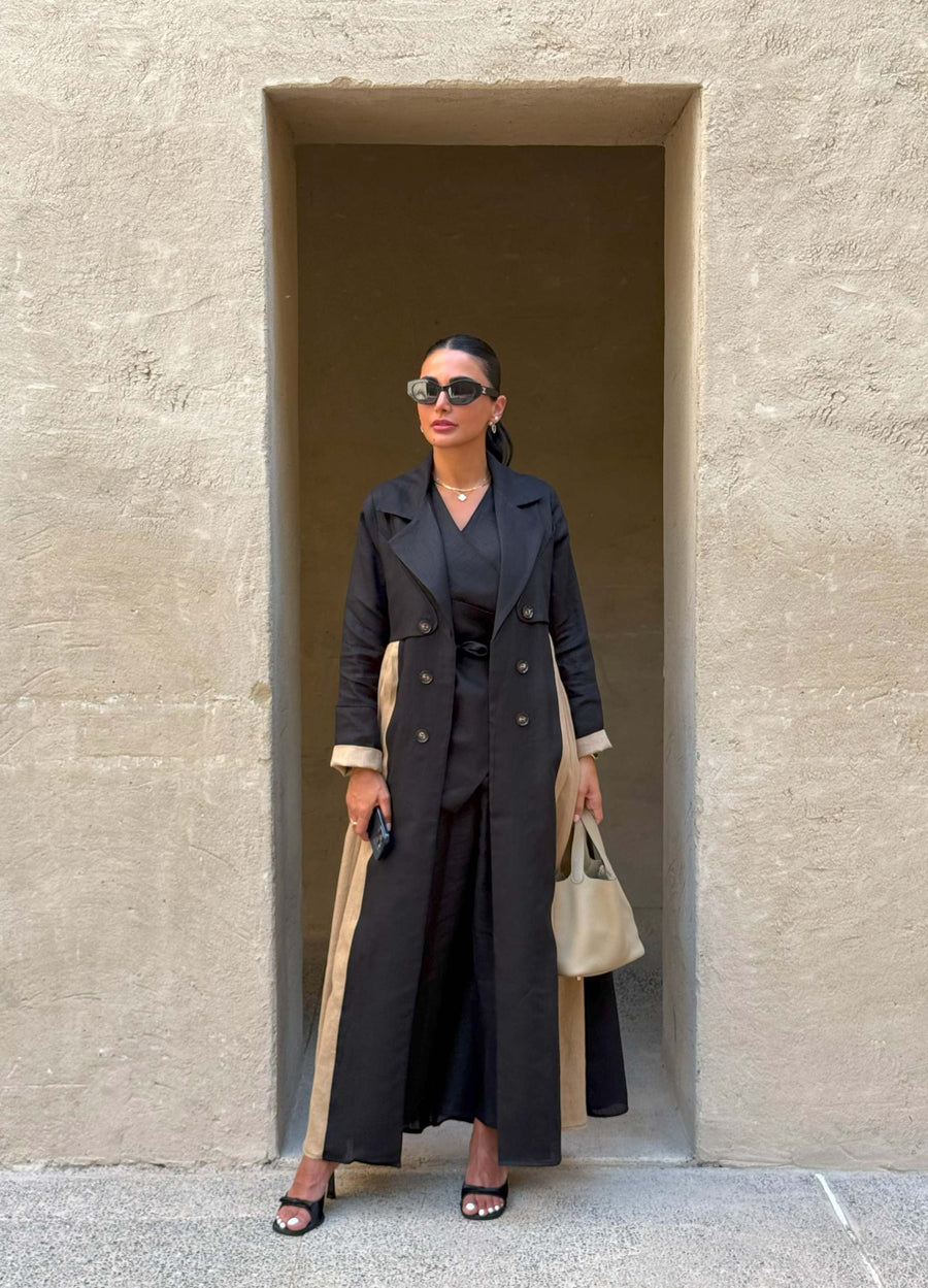 Woman wearing Latte Smoke trench-style black abaya with beige side panels and cuffs, double-breasted design and structured collar, holding beige handbag.