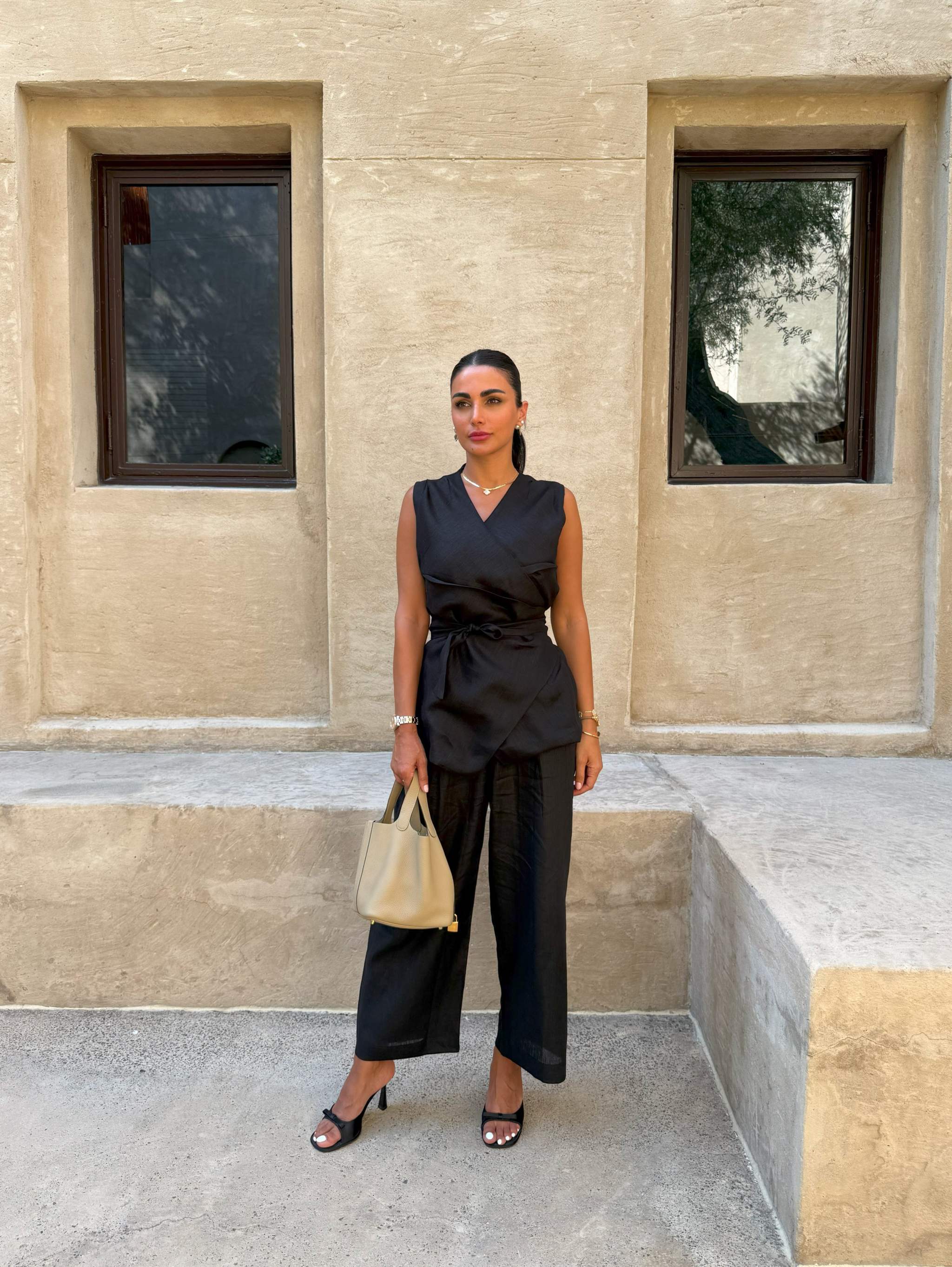 Black sleeveless Chai Linen Vest with wrap design and self-tie belt, styled with black pants and beige handbag.