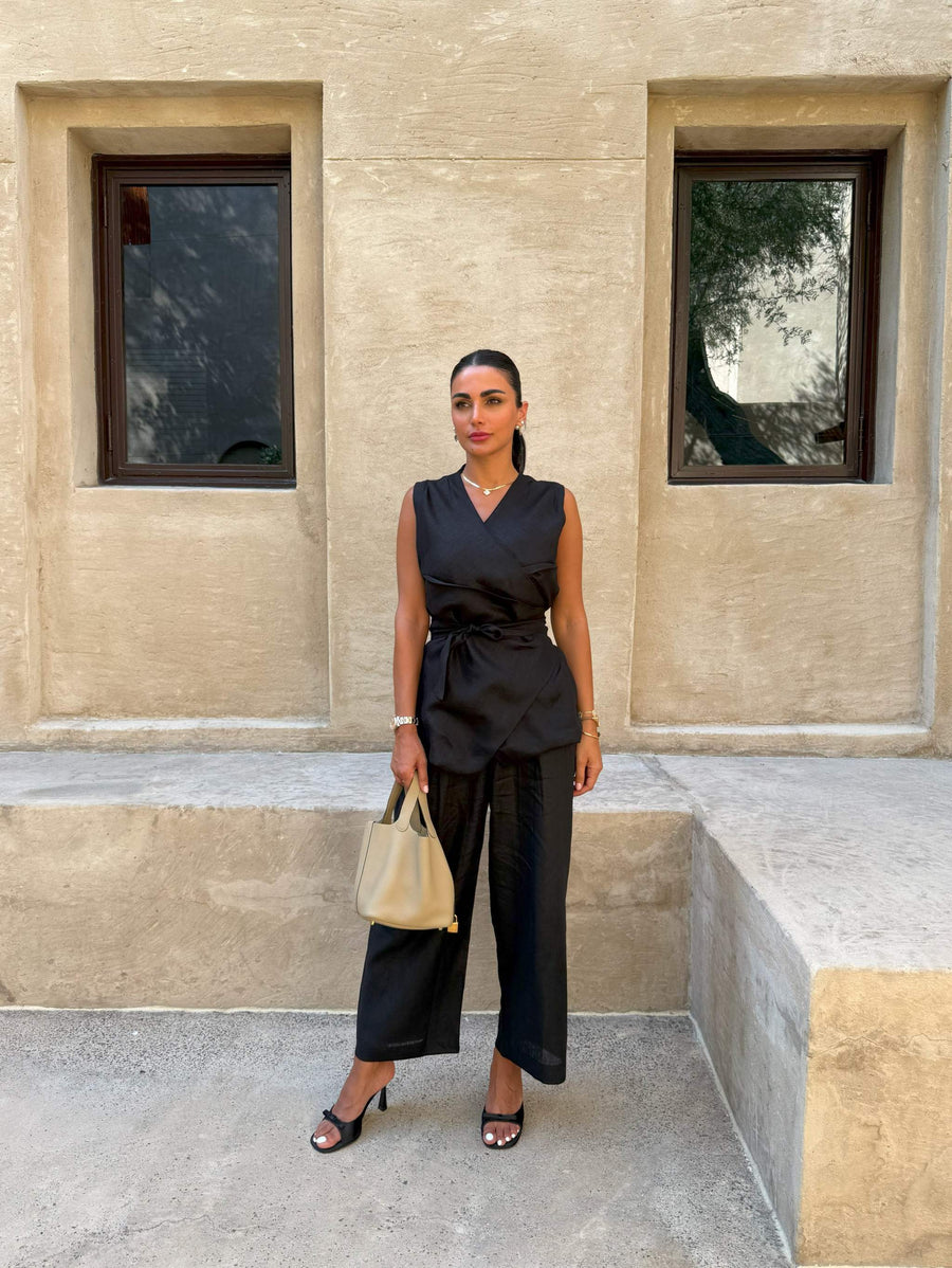 Black sleeveless Chai Linen Vest with wrap design and self-tie belt, styled with black pants and beige handbag.