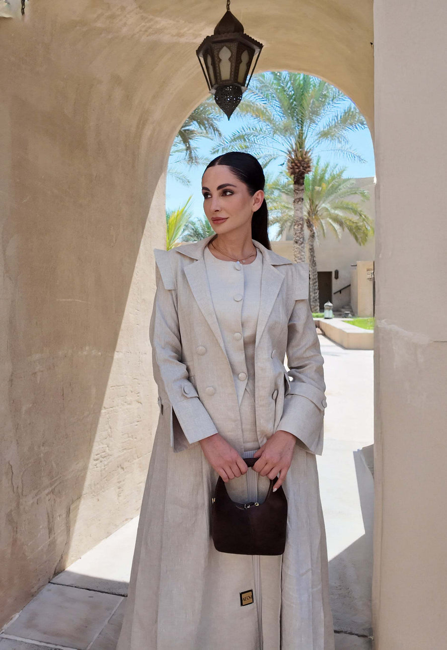 woman wearing a beige linen abaya with trench-style silhouette and matching sheila holding a brown handbag - beige abaya