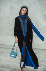 Woman wearing elegant black and blue velvet abaya with blue accents and matching scarf carrying blue handbag