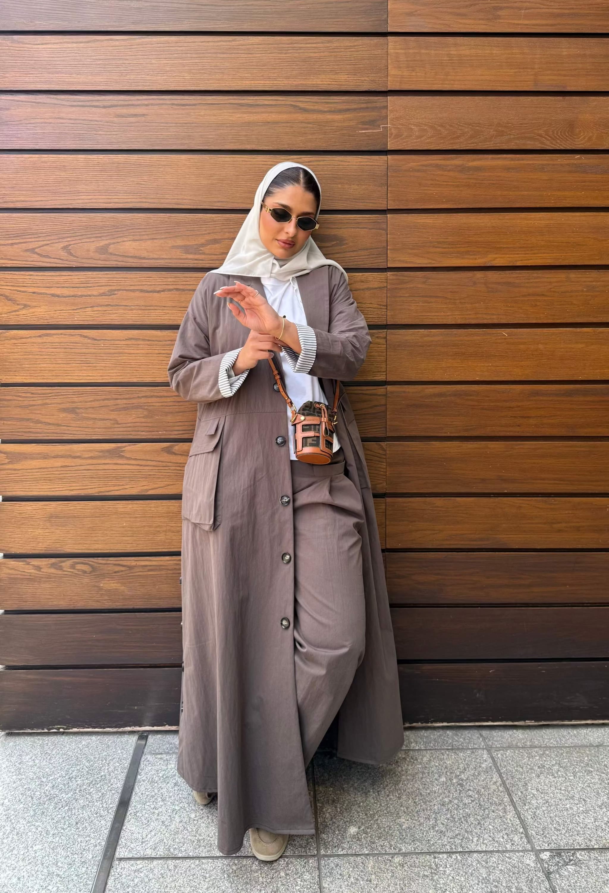 Woman wearing taupe wide-leg pants with a matching long coat and white hijab standing against a wooden wall